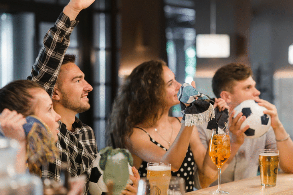 Grupo de personas animando y viendo un partido de fútbol en un bar mientras disfrutan de bebidas, representando la emisión de televisión de pago y la importancia de cumplir con los derechos de autor en hostelería en España.