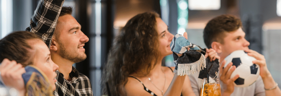 Grupo de personas animando y viendo un partido de fútbol en un bar mientras disfrutan de bebidas, representando la emisión de televisión de pago y la importancia de cumplir con los derechos de autor en hostelería en España.